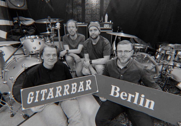 En svartvit bild på fyra killar framför ett trumset, hållandes i två skyltar där det står Gitarrbar Berlin.