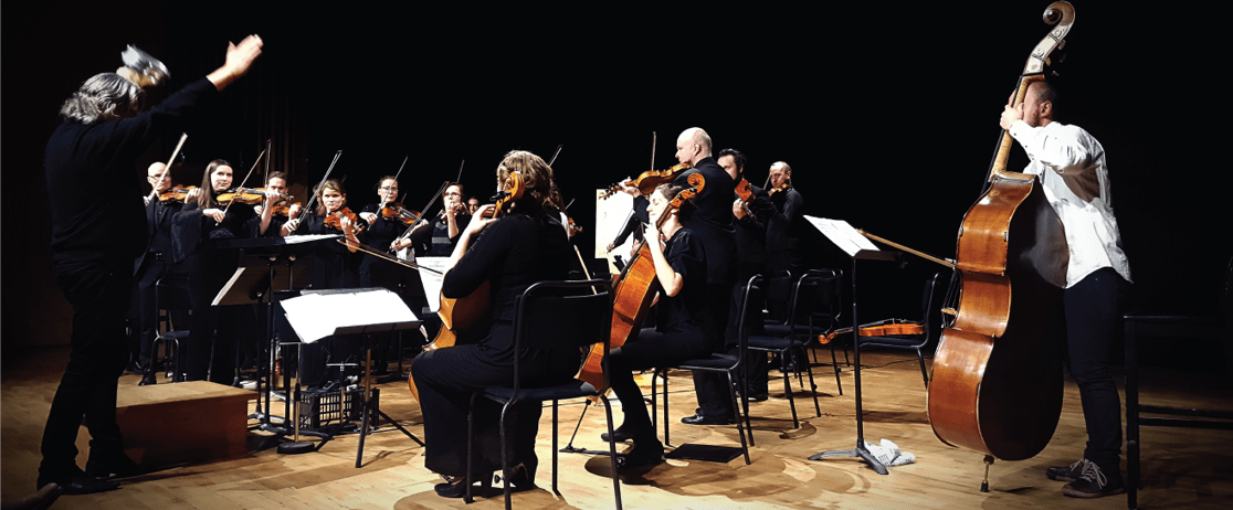 Dalslandsorkesterns stråkmusiker blir dirigerade på en scen mot en svart bakgrund.