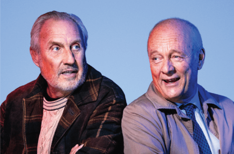 De två skådespelarna sitter i vardagliga kläder mot en blå bakgrund