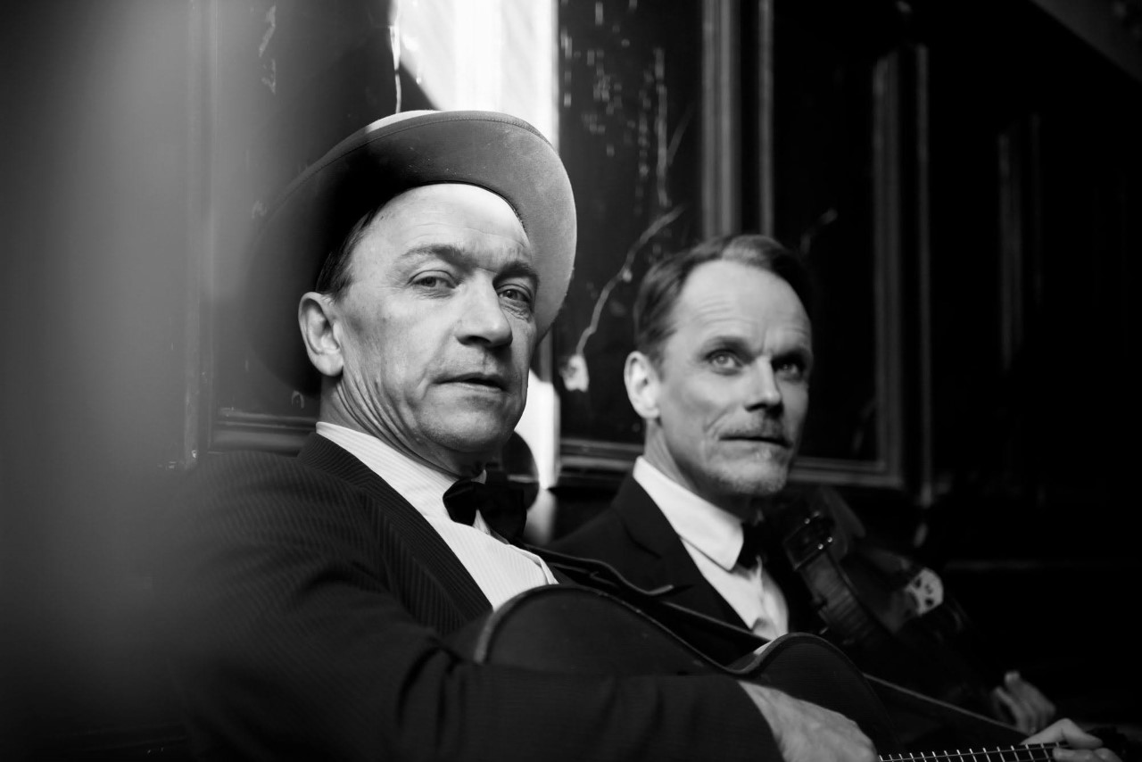 Schlagerkavaljererna Erik Drougge och Magnus Marcinkowski Pettersson är fotograferade från sidan, de håller i en gitarr respektive en fiol. Det är ett svartvitt foto.