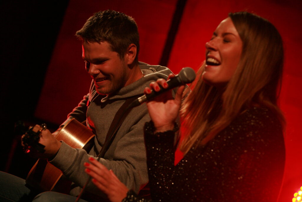 Emil Ernebro spelar gitarr och Zandra Ernebro sjunger på en rött upplyst scen. De ser glada ut.