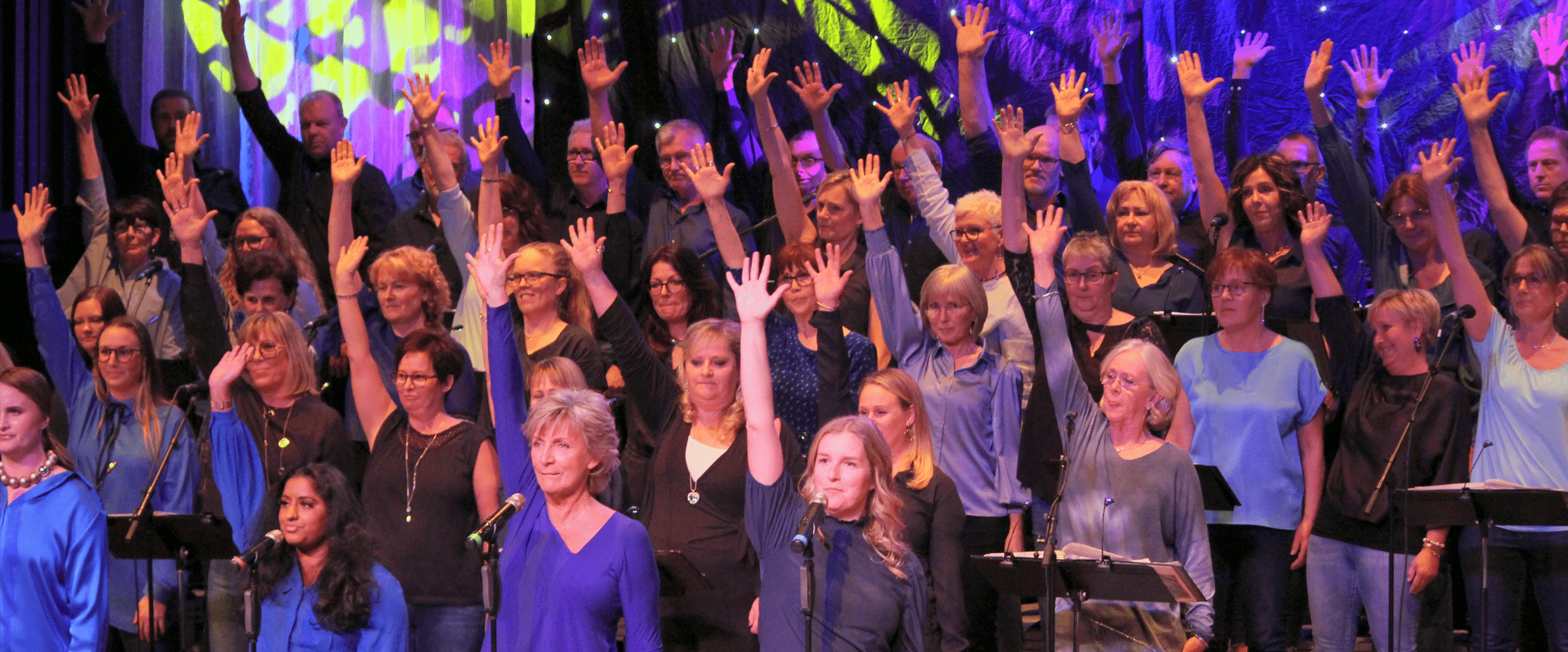 Kören Song of Joy sjunger på en blå-lila upplyst scen.