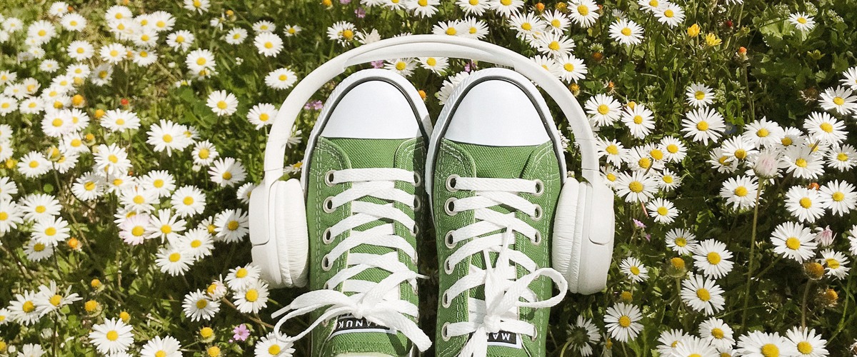 Ett par gröna skor vilar på en gräsmatta med tusenskönor, runt skorna sitter ett par vita hörlurar.