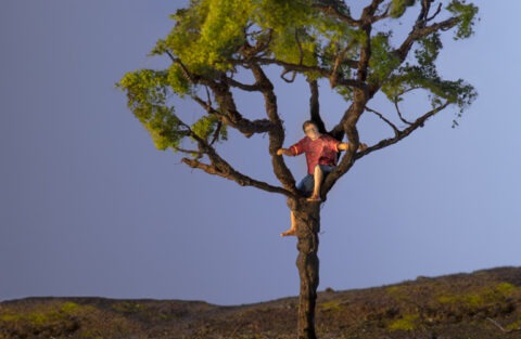 En konstnärligt tillverkad pojke sitter uppklättrad i ett träd mot en blå himmel.