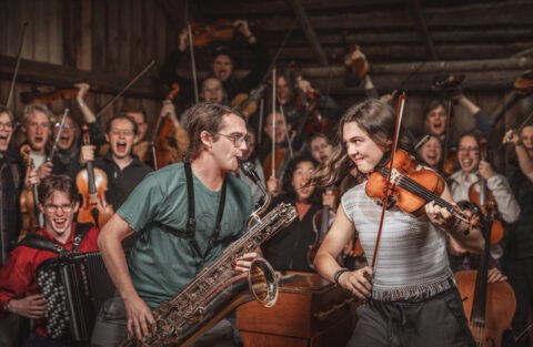 En saxofonist och en violinist spelar och tittar varann i ögonen, bakom står massor av glada musikanter.
