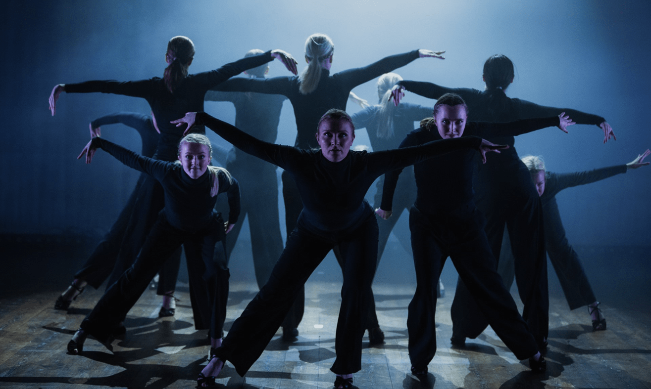 Tio dansare klädda i svart står på en rökig, blålyst scen i dramatiska positioner.