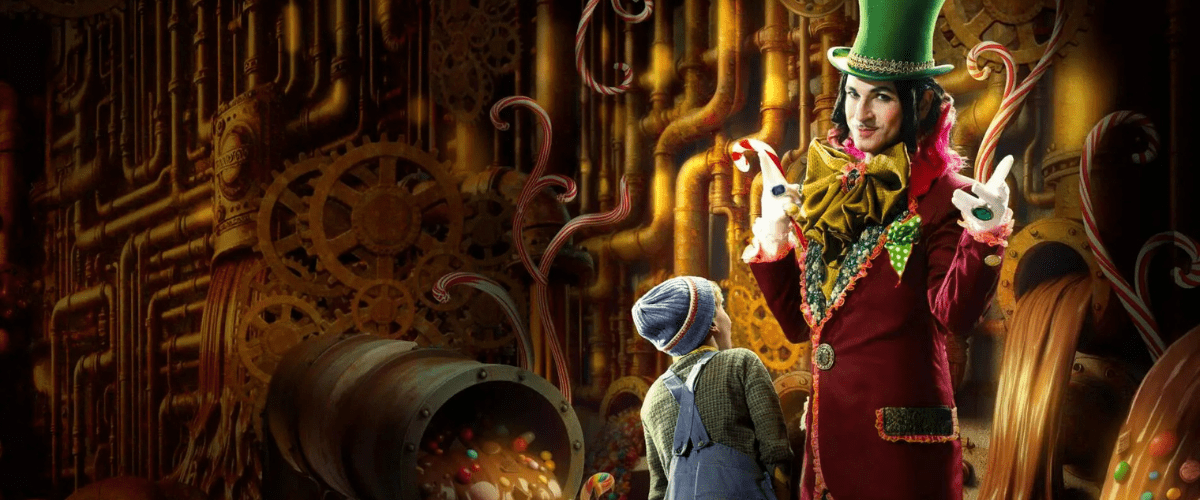 Willy Wonka tittar in i kameran framför en vägg av rör. Bredvid honom står Kalle i blå hängslebyxor och mössa.