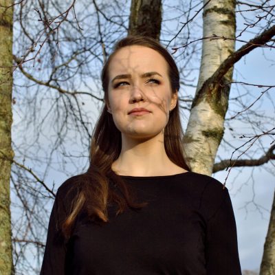 Victoria Alice tittar åt sidan med ett litet leende, stående framför några björkar.