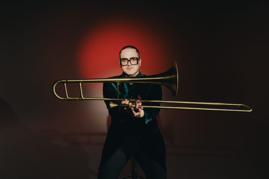 Matteo Paggi håller i sin trombon framför en röd vägg. Han har markerade, stora glasögon och en kavaj i grön, krossad sammet.