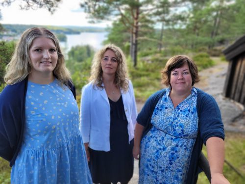 Arvejols tre medlemmar klädda i blått framför en vacker utsikt med vatten, klippor och träd.