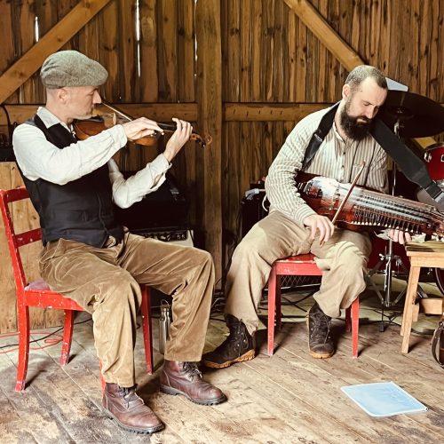 Två män på en varsin röd trästol i en lada. En spelar fiol, en spelar nyckelharpa.