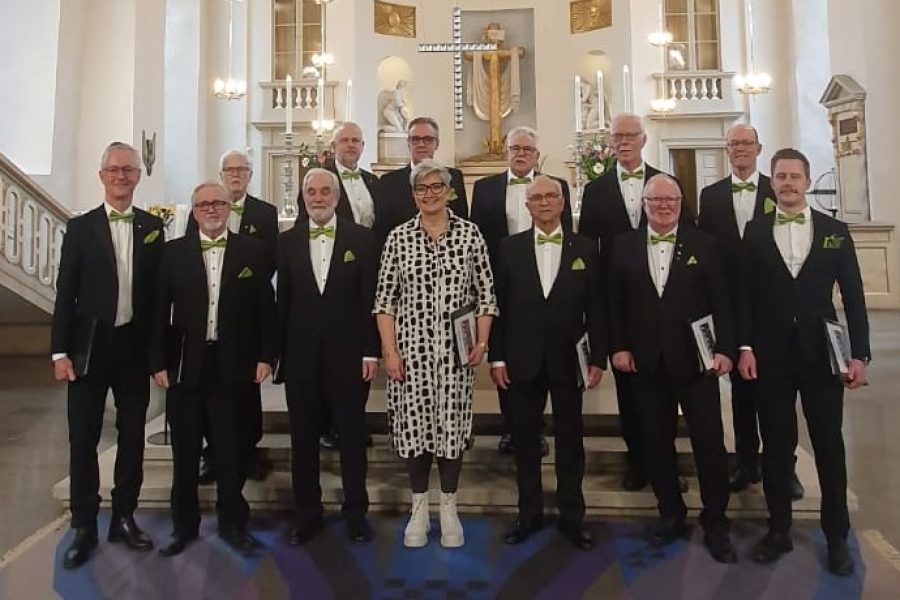 Kören Bettes Drängar står uppställda i kostym, tillsammans med sin ledare i svartvit klänning, framför ett altare.