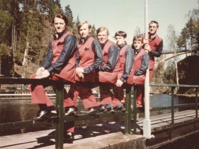 En gammal bild på Jimsons när de sitter iklädda röda och svarta kostymer på ett kanalräcke.