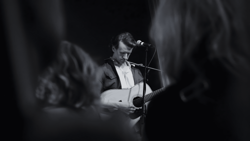 Martin står på en scen och skymtas spelandes gitarr mellan publikdeltagare.