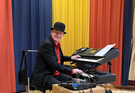 Robert i svart kostym, plommonstop och röd skjorta vid två keyboards och ett mixerbord.