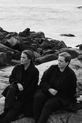Två kvinnor sitter på en klippa vid havet, det är en svartvit bild.