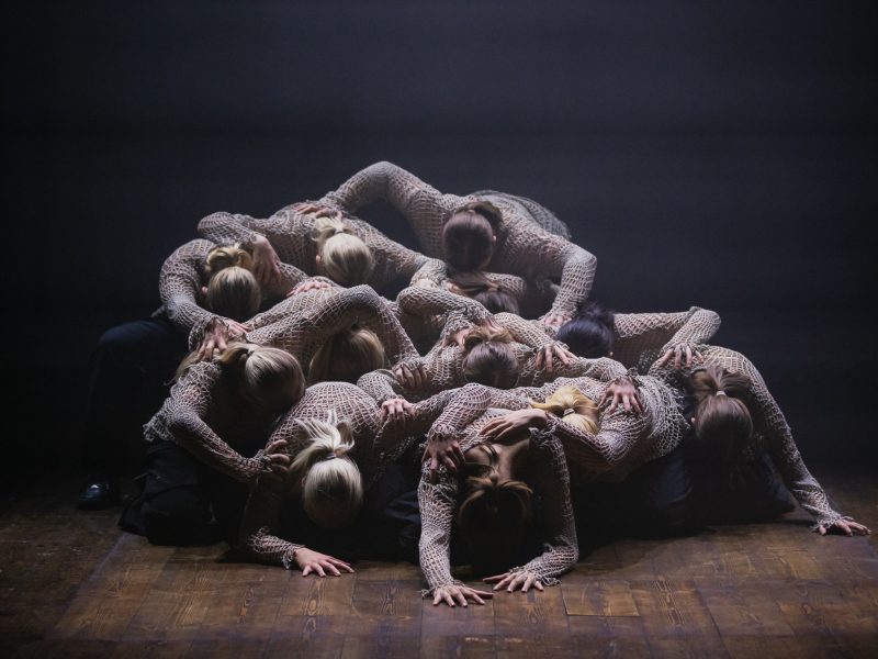Elva dansare ligger i en hög iklädda bruna nättröjor på ett scengolv.