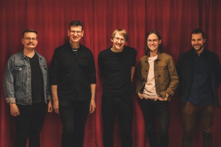 chris erik band De fem medlemmarna i Chris Erik Band står framför ett rött draperi och skrattar in i kameran.