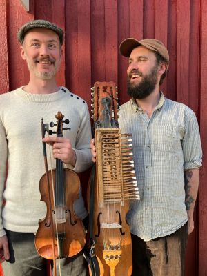 Tom Cubbin med nyckelharpa och Alex Rawlins med fiol står framför en röd husvägg och ler.