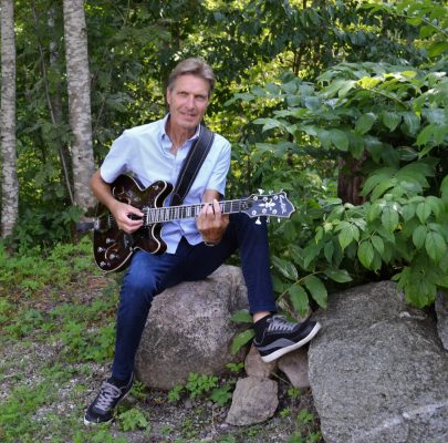 Stig-Arne Larsson sitter på en sten framför en buske och spelar elgitarr.